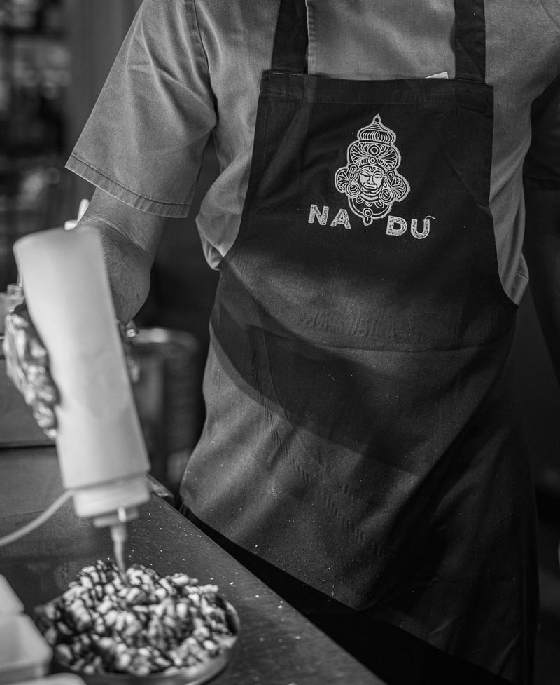 A member of the Nadu team in a branded apron, finishing a dish at the pass